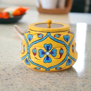 Puede incluir: Un azucarero de cerámica amarilla con tapa y una cuchara pequeña. El cuenco está decorado con motivos florales azules y un borde azul. La tapa tiene un pequeño pomo de madera. El texto "Gauri Arts India" está impreso en la tapa.