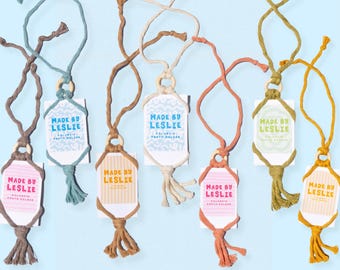 May include: A collection of macrame photo holders in various colors including brown, blue, tan, cream, orange, green, and yellow. Each holder features a wooden ring, a hanging cord, and a card with the text Made By Leslie Polaroid Photo Holder.