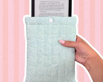 May include: A light mint green quilted fabric sleeve featuring a repeating white smiley face pattern. The rectangular pouch has a single white snap button closure at the top center and contains a black e-reader displaying visible text.