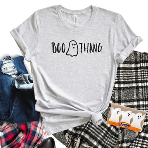 May include: A gray t-shirt with the text "BOO THANG" and a ghost graphic. The shirt is tied in a knot and is laying on a black and white plaid blanket. A box of white Peeps marshmallow candies is also on the blanket.