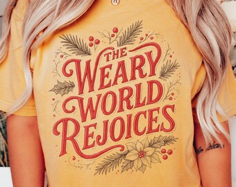 May include: Mustard-colored t-shirt with the text "The Weary World Rejoices" in red script. The design includes festive botanical elements like holly, berries, and pine needles. The shirt is a comfortable, casual style.