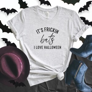 May include: Light gray t-shirt with the text "IT'S FRICKIN bats I LOVE HALLOWEEN" in black. The shirt is styled with a burgundy hat, black boots, and bat silhouettes.