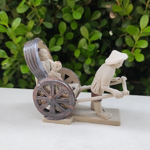 May include: A ceramic figurine of a person pulling a rickshaw with another person sitting inside. The rickshaw is brown and the figures are beige.