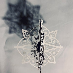 May include: A white paper snowflake ornament with intricate cutouts hangs from a string. The snowflake is suspended in front of a blurry background, casting a shadow.