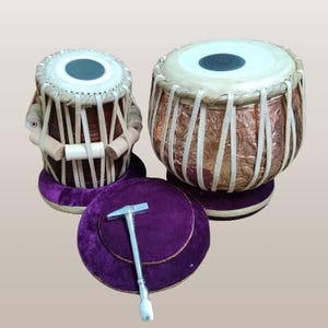 Puede incluir: Un conjunto de dos tambores tabla marrones y de cobre con cojines de terciopelo morado y un martillo de afinación plateado. Los tambores son instrumentos de percusión tradicionales de la India.