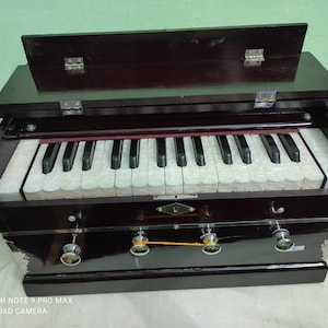 May include: A dark brown wooden harmonium with a keyboard and bellows. The keyboard has white and black keys and the bellows have a decorative design.