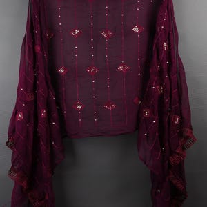 May include: A sheer, plum-colored scarf with a pattern of diamond shapes and small, scattered sequins. The scarf has a fringed edge and is draped against a gray background. The fabric appears lightweight and flowing, suitable for layering.