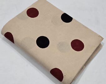 May include: Folded beige fabric with large, evenly spaced polka dots in black and burgundy. The fabric is folded over itself, showing the pattern on the top layer. The fabric appears to be cotton or a similar material.