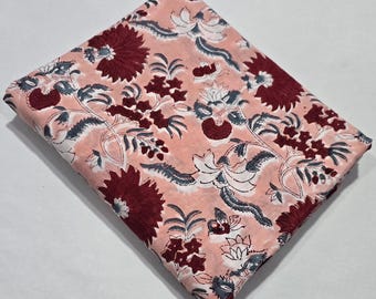 May include: Folded fabric with a floral pattern. The fabric features a light peach background with a repeating design of burgundy flowers, white blossoms, and gray-blue leaves and stems. The fabric is folded and ready for use.