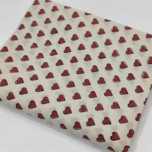 Puede incluir: Un trozo de tela doblado con un patrón repetido de corazones rojos sobre un fondo crema. Cada corazón está delineado en negro. La tela parece ser de algodón o un material similar, adecuado para proyectos de costura o manualidades.