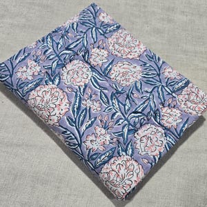 Pode incluir: Tecido dobrado com um padrão floral. O design apresenta grandes flores rosa e brancas, folhas e caules azuis sobre um fundo lavanda. O tecido é provavelmente algodão, ideal para projetos de patchwork ou artesanato.
