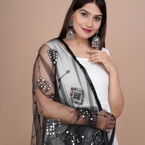May include: A black net dupatta with mirror embellishments. The dupatta features a square pattern of mirrors and is draped over a white dress. Silver-toned earrings with a bell design are also visible.