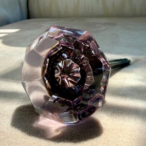 May include: A faceted, octagonal glass door knob with a light purple tint. The knob features a central, star-shaped indentation and a metallic spindle extending from the back. The glass surface is transparent with geometric edges and facets.