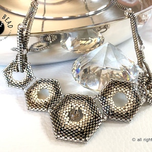 May include: A silver necklace with a geometric design featuring three large hexagonal beads with open centers. The necklace is on a white surface with a clear crystal next to it.