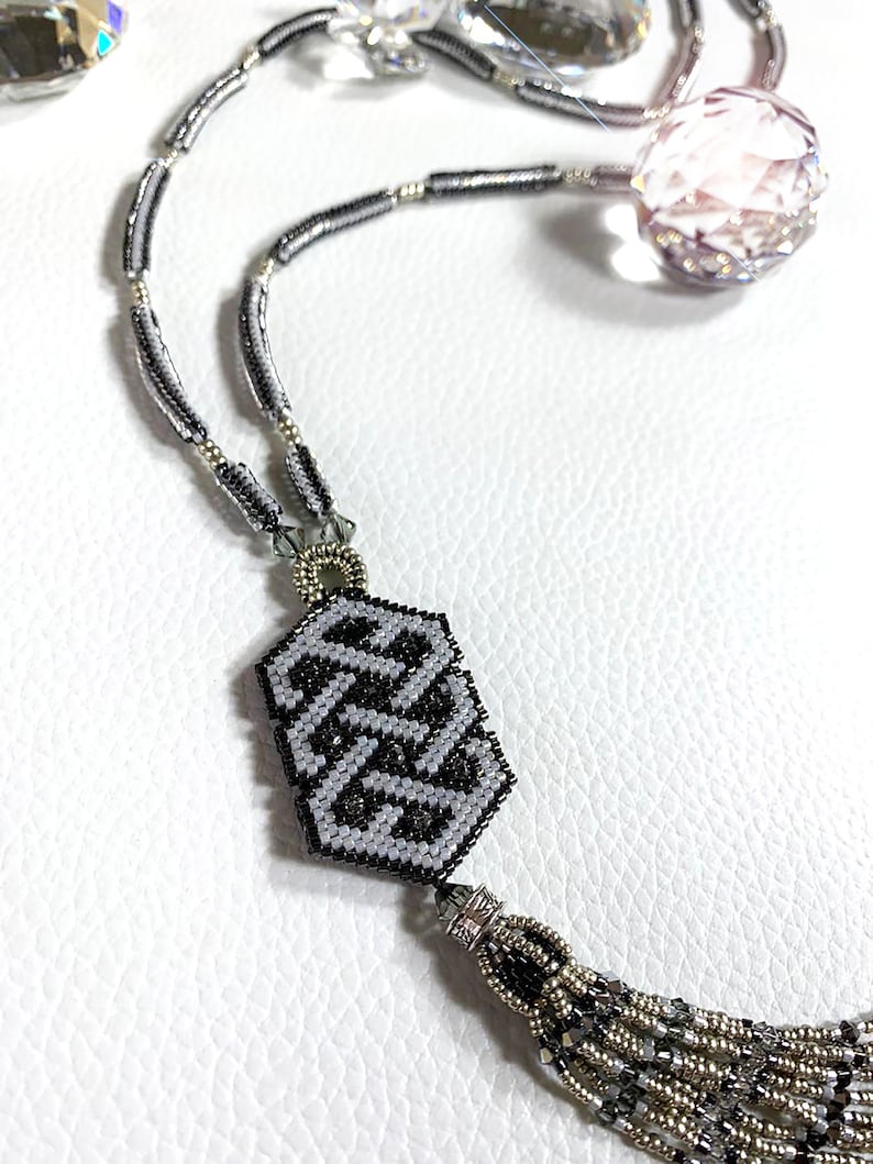 May include: A silver and black beaded necklace with a geometric pendant and a tassel. The pendant is made of black and white beads and has a geometric pattern. The tassel is made of silver and black beads.