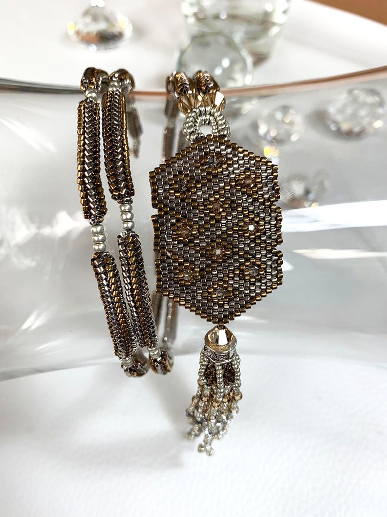 May include: A pair of beaded bracelets with a silver and gold color scheme. The bracelets are made of small, round beads and have a geometric pattern. The bracelets are accented with a tassel.