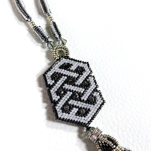 May include: A black and white beaded necklace with a geometric pendant and a tassel. The pendant is made of small black and white beads and has a geometric pattern. The tassel is made of black, white, and silver beads.