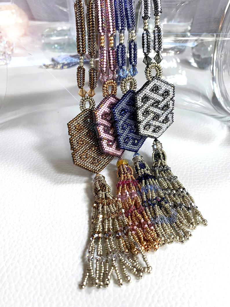May include: Five beaded necklaces with hexagonal pendants and tassel ends. The pendants are in shades of gold, pink, blue, and gray. The tassels are in shades of gold, pink, blue, and silver.