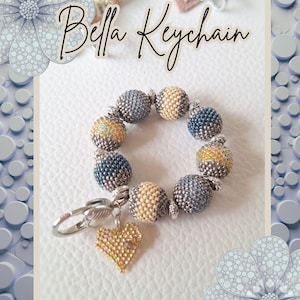 May include: A beaded keychain with the text "Bella Keychain". The keychain features a ring of beaded spheres in shades of blue, beige, and gold. A gold heart charm hangs from the silver clasp.