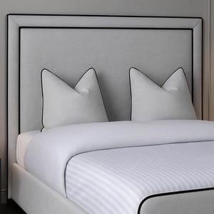May include: A contemporary bed featuring a grey upholstered headboard with black trim. Two white pillows with black piping are placed against the headboard. The bed is made with a white striped duvet cover and a white sheet.