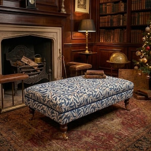 May include: A rectangular ottoman upholstered in a blue and cream fabric featuring a repeating leaf and branch pattern. The piece is supported by four dark wooden legs with small metal casters, and it is topped with two stacked vintage books.