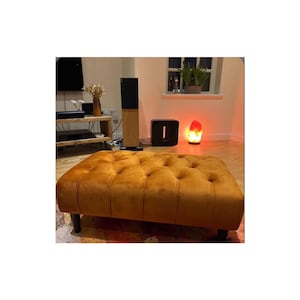 May include: A mustard yellow velvet tufted ottoman with wooden legs. The ottoman is in a living room setting with a salt lamp and a speaker in the background.