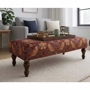 May include: A rectangular ottoman upholstered in a deep red fabric featuring a repeating pattern of golden birds and floral motifs. The piece is supported by four dark brown turned wooden legs and holds a dark wood tray with books and an hourglass.