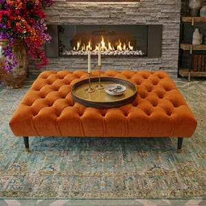 May include: A burnt orange, button-tufted ottoman with a decorative tray, candles, and a small bowl. The ottoman is rectangular and rests on black legs with gold detailing. The background features a fireplace and a patterned rug.