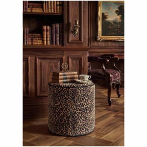 May include: A cylindrical ottoman upholstered in a brown and black leopard print fabric with black piping along the top and bottom edges. The stool features a firm, flat top surface and a structured, circular base design for stability.