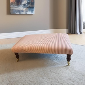 May include: A square ottoman with a red and white striped fabric top. The ottoman has dark wooden legs with brass caster wheels. The piece is set on a light grey rug, suggesting a living room or bedroom setting.