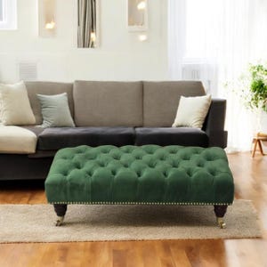 May include: An emerald green, button-tufted ottoman with nailhead trim. The rectangular ottoman has dark wooden legs with brass castors. A neutral rug and a grey sofa are in the background.