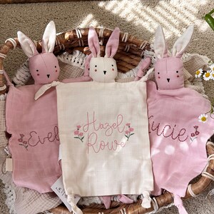 May include: Three pink bunny lovey security blankets with embroidered names "Evel", "Hazel Rowe", and "Lucie". Each bunny has a white face and long ears. The blankets are in a woven basket.