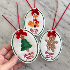 Può includere: Tre ornamenti ovali in feltro bianco con bordi verdi smerlati e fiocchi di nastro rosso. Uno presenta una volpe ricamata, uno un albero di Natale verde e uno un omino di pan di zenzero. Ognuno è personalizzato con un nome e l'anno 2026.