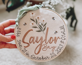 May include: A beige embroidered hoop with the name "Sayler Bay" in brown cursive lettering. The hoop also includes the date "October 25, 2023" and the text "8 lbs & 21 in" in black lettering. The hoop is decorated with a floral design and small embroidered bees.