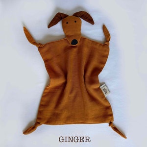May include: A brown muslin baby security blanket with a brown felt dog face and black felt eyes and nose. The blanket has knotted corners and the text "GINGER" is printed below the blanket.