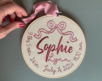 May include: A 7-inch embroidery hoop with a pink ribbon and the name "Sophie Lynn" embroidered in red thread. The hoop also includes the date "July 14, 2024" and the time "9:55 pm 1:30 am".
