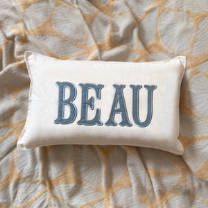 May include: A cream-coloured rectangular cushion featuring the word "BEAU" embroidered in a blue, decorative font. The cushion rests on a patterned beige and white fabric, creating a soft and inviting aesthetic.