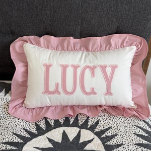 May include: A white rectangular pillow with the name "LUCY" embroidered in pink, framed by a pink ruffled border. The pillow rests on a patterned quilt with a black and white sunburst design.