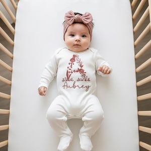 May include: A baby wearing a white onesie with a floral "A" design and the name "Aubrey" in brown script. The baby is also wearing a mauve bow headband. The baby is lying in a wooden crib with a white mattress.