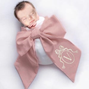 May include: A pink bow-shaped baby swaddle with a white embroidered monogram of the letter "V" in a decorative frame. The swaddle is wrapped around a sleeping baby.
