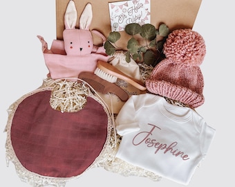May include: A pink and white baby gift box with a pink bunny, a pink knitted hat, a white onesie with the name Josephine embroidered on it, a wooden hair brush, a comb, and a pink bib.