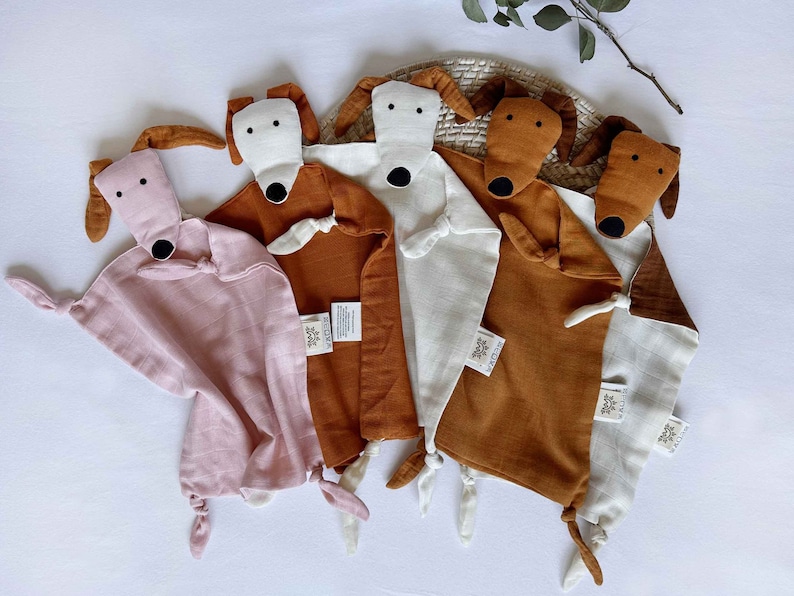 May include: Four plush dog-shaped baby security blankets. The blankets are made of soft, muslin fabric and come in pink, white, and two shades of brown. Each blanket features a cute dog face with black eyes and a black nose.