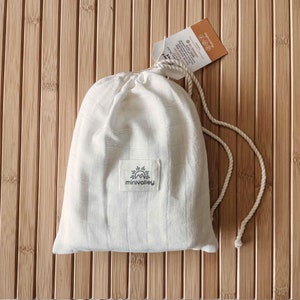May include: White muslin swaddle with a drawstring closure and a small black logo that says "minivalley" with a sun and mountain design. The swaddle is on a wooden surface.