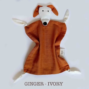 May include: A soft, brown and ivory plush dog toy with a white face and black nose. The toy has four white ties attached to the corners.