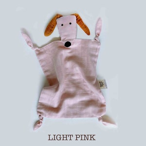 May include: A light pink muslin baby security blanket with a brown and black dog face. The blanket has knotted corners and a small white tag with the word "BESTOW" on it.
