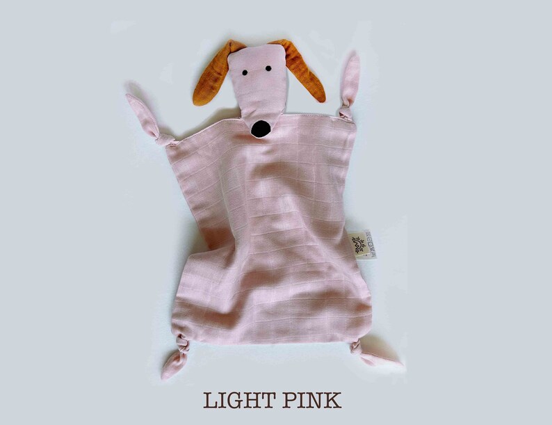 May include: A light pink muslin baby security blanket with a brown and black dog face. The blanket has knotted corners and a small white tag with the word "BESTOW" on it.