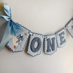 May include: A decorative banner featuring scalloped fabric panels with a light blue and white gingham pattern. The panels display an embroidered white goose with a blue bow and the letters O, N, and E in dark blue. A light blue satin ribbon bow.