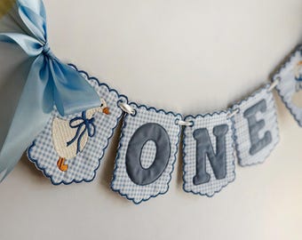 May include: A decorative banner featuring scalloped fabric panels with a light blue and white gingham pattern. The panels display an embroidered white goose with a blue bow and the letters O, N, and E in dark blue. A light blue satin ribbon bow.