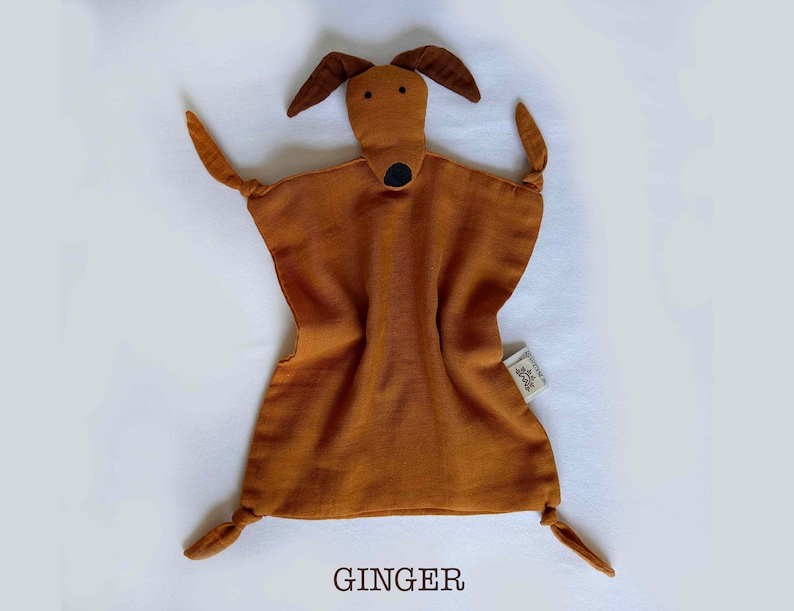 May include: A brown muslin baby security blanket with a brown felt dog face and black felt eyes and nose. The blanket has knotted corners and the text "GINGER" is printed below the blanket.