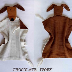 May include: A brown and white muslin baby security blanket with a brown dog head attached. The blanket has a white muslin center and brown muslin edges. The dog head has a brown nose and black eyes. The blanket has a tag that says "VEDUTA".